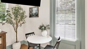 A dining area with a round table, two chairs, a potted plant, a framed picture, and a window with blinds. - "Idlehour" new cottage in Fairhope, AL - NEW LISTING (Fairhope)