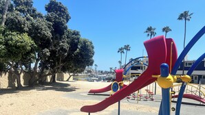 Une aire de jeux avec un toboggan rouge et des structures bleues et jaunes, entourée d’arbres et d’une zone sablonneuse.