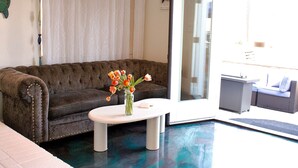 Un salon avec un canapé marron à motifs capitonnés, une table basse blanche ornée d’un vase de fleurs et une porte vitrée donnant sur un espace extérieur où se trouve un canapé en osier.