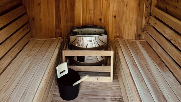 Una sauna con paredes y bancos de madera, un banco de madera en el centro y un cubo negro con asa blanca.