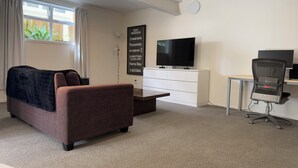 Una habitación de hotel moderna con un sofá marrón, un televisor de pantalla plana, un escritorio con silla y un ventanal con cortinas.