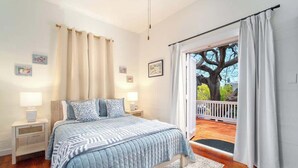 A bedroom with a large bed, a nightstand, a window with curtains, and a view of a tree outside. - Key Lime Hideaway (Key West)