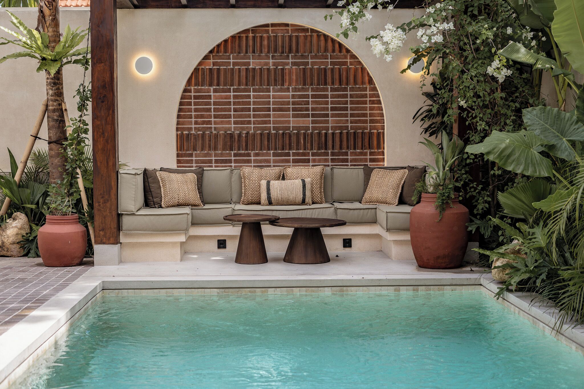 Un área de descanso junto a la piscina con un sofá y dos mesas redondas, rodeada de plantas en macetas y un muro de ladrillo con un ventanal circular.
