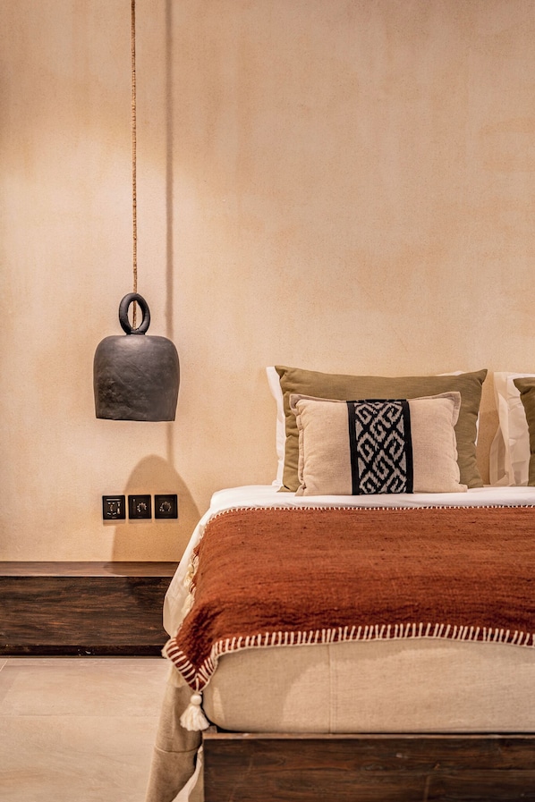 Una cama bien tendida con un edredón marrón y una almohada estampada, una lámpara colgante y un reloj fijado a la pared.