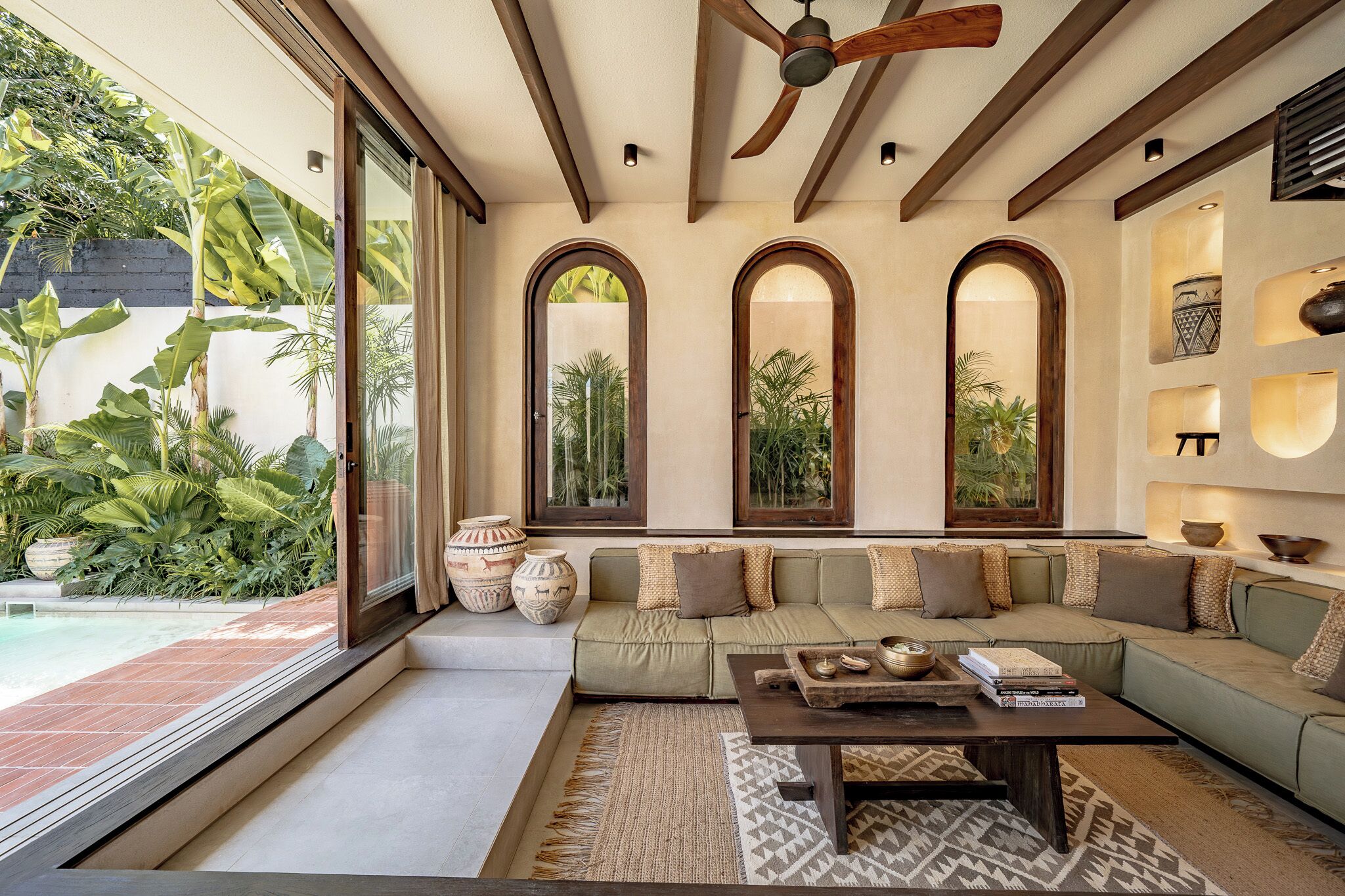A modern living room with a green sofa, a coffee table, and a ceiling fan. There are large windows with arched tops and a view of greenery outside.