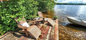 A wooden dock with two Adirondack chairs and a small table, overlooking a calm lake with a boat moored nearby. - Island Cabin + Glamping for 5 | Boats + Hot Tub (Greig)