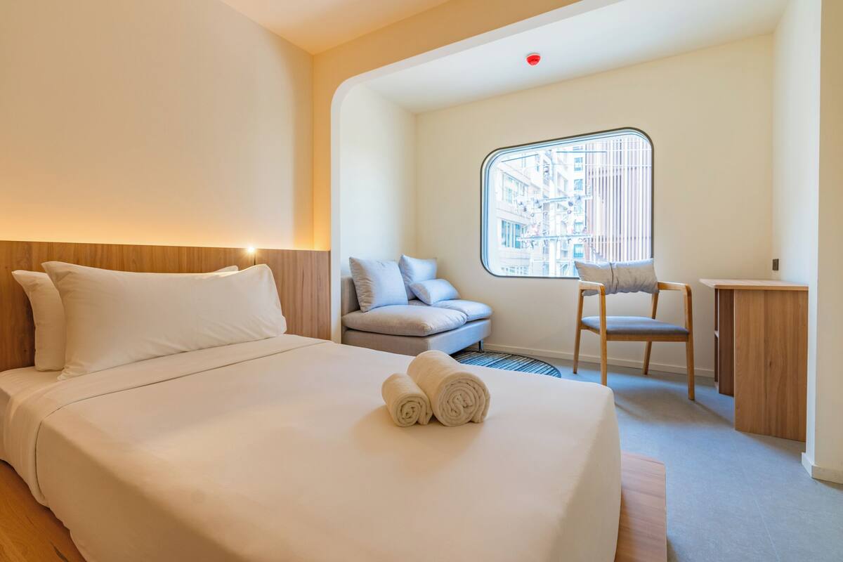 Ein Hotelzimmer mit einem großen Bett, einem kleinen Schreibtisch, einem Sessel und einem Fenster mit Blick auf die Stadt.