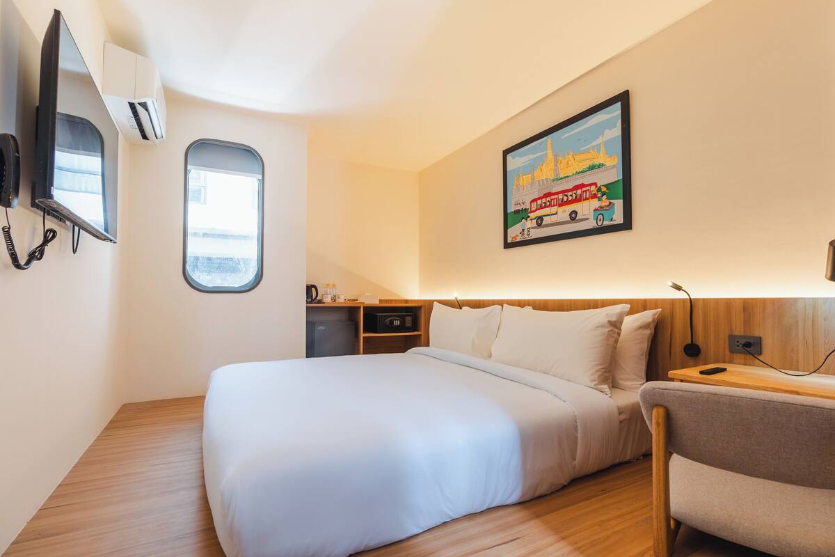 Ein Hotelzimmer mit einem Bett, einem kleinen Schreibtisch, einem Stuhl, einem Fenster, einer Klimaanlage, einem Telefon, einem Gemälde und einem Nachttisch mit einer Lampe.