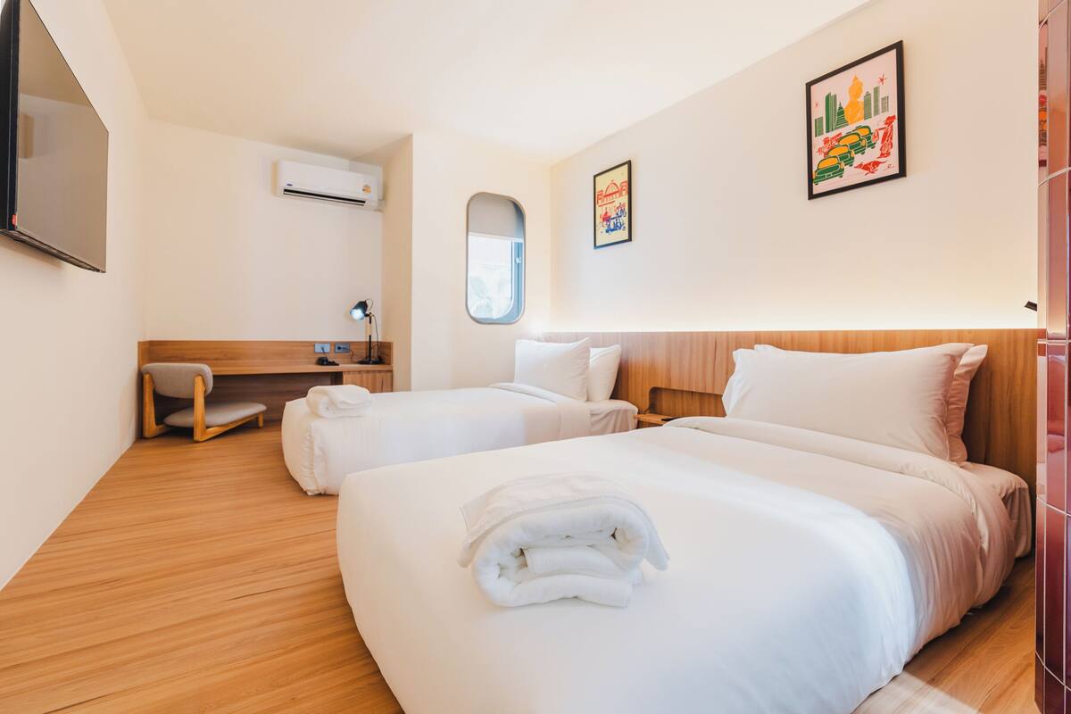 Ein Hotelzimmer mit zwei Betten, einem Schreibtisch, einem Sessel, einer Klimaanlage, einem Fenster und einem gerahmten Bild an der Wand.