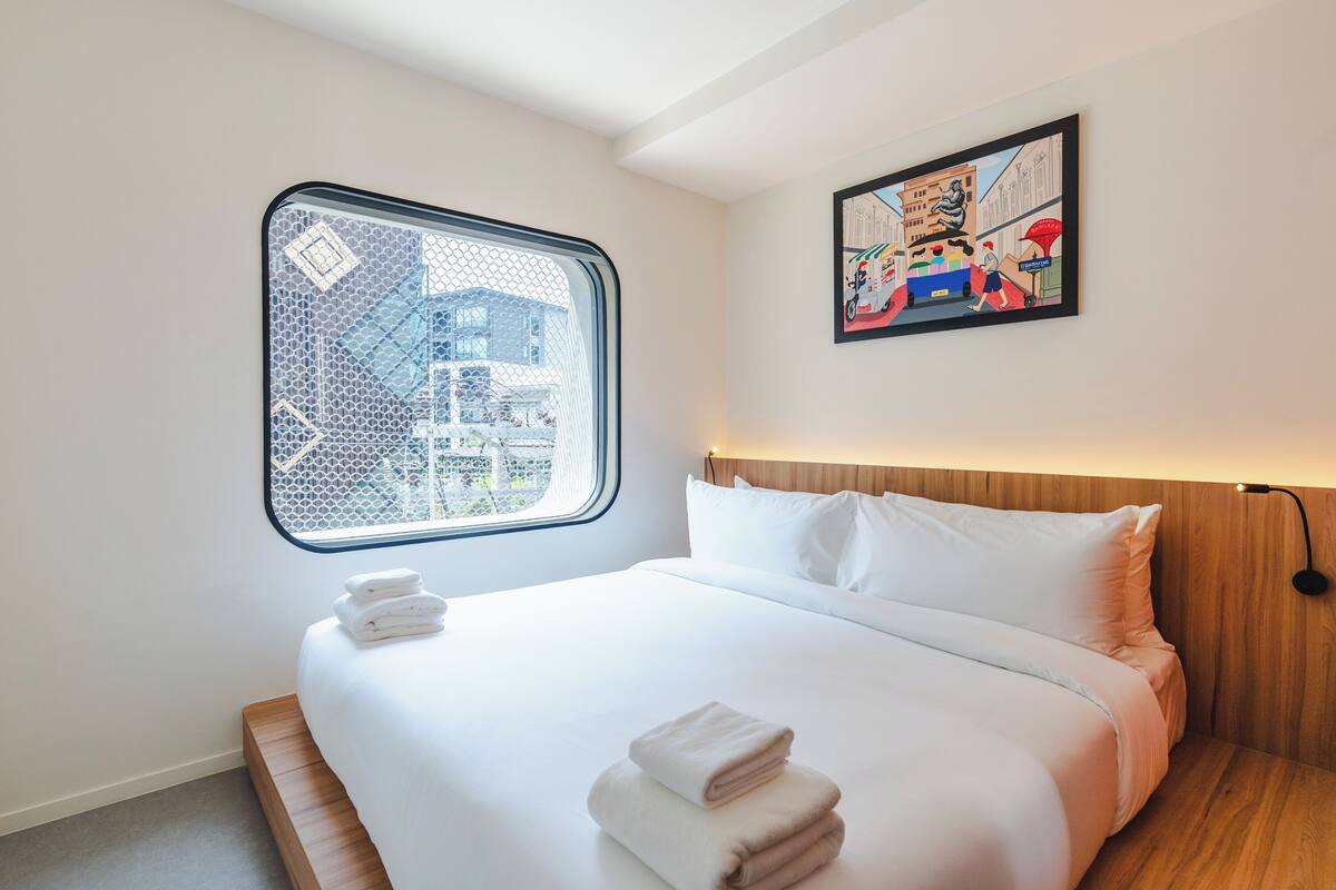 Ein Hotelzimmer mit großem Fenster, einem Bett mit weißer Bettwäsche und einem farbenfrohen Kunstwerk an der Wand.