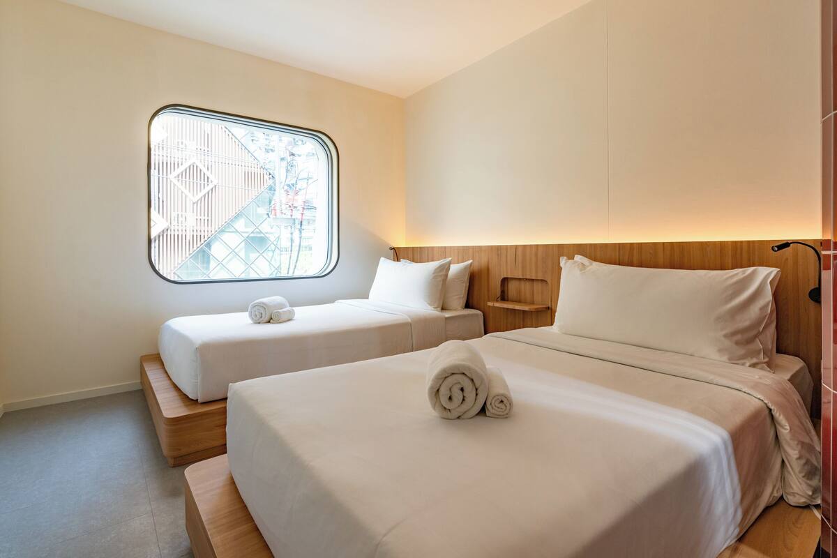 Ein Hotelzimmer mit zwei Betten, einem Fenster mit Blick auf Gebäude und einem hölzernen Kopfteil.
