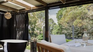 A covered outdoor seating area with a black bathtub, a table with a bottle, and a view of trees. - A Romantic Escape in Nature’s Embrace (Bullarto South)
