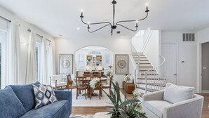 Ein geräumiger Wohnbereich mit blauem Sofa, grauem Sessel, Esstisch mit Stühlen, Treppe und einer Chandelieber.