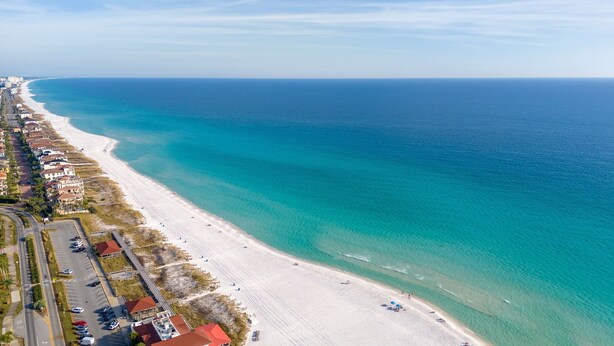 Eine Luftaufnahme einer Küstenlandschaft mit einem langen Sandstrand, klarem blauen Wasser und einer Reihe von Gebäuden entlang der Uferlinie.