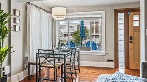 Una habitación con una mesa de comedor y sillas, una ventana que da a vistas de casas, una puerta y una planta en maceta.