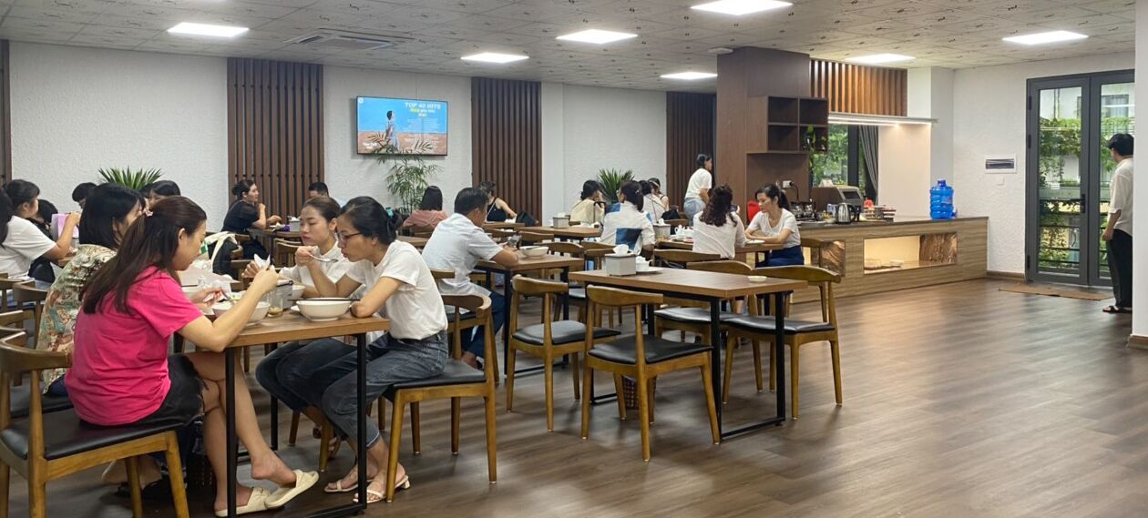 A dining area with wooden tables and chairs, a counter with a water dispenser, and a large screen displaying a presentation.