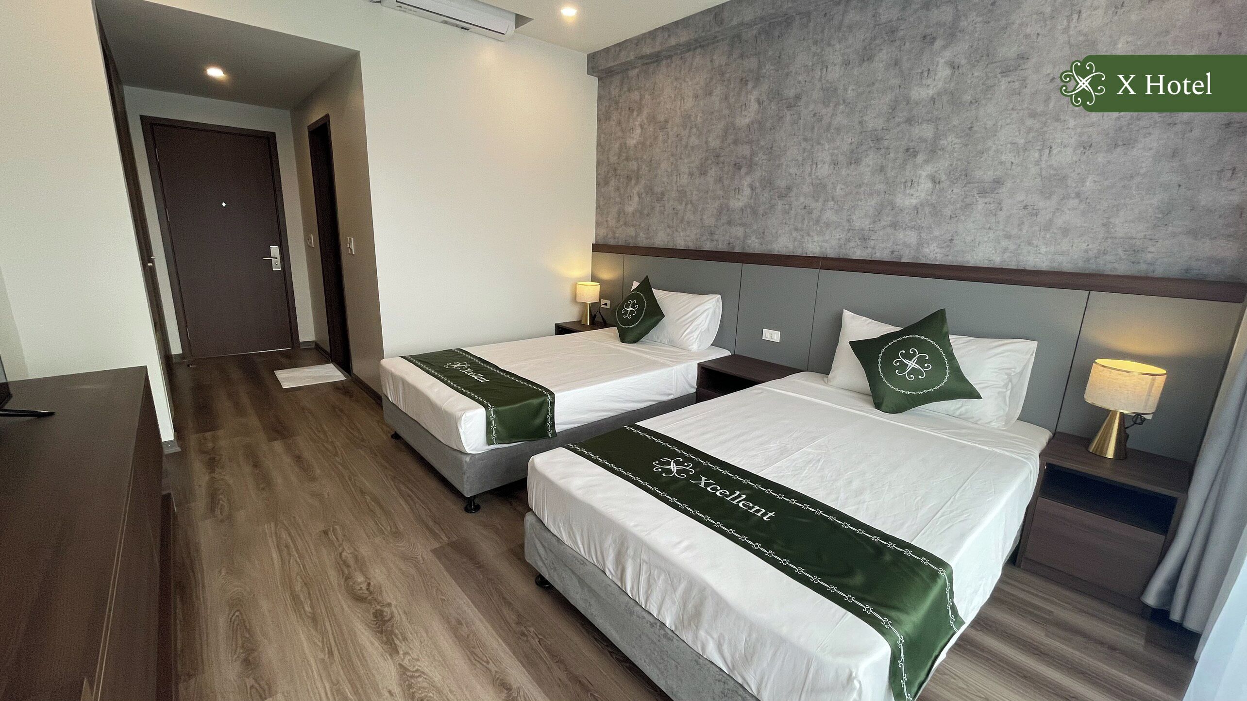 Habitació d'hotel amb dos llits, un sòl de fusta i una paret gris texturitzada.