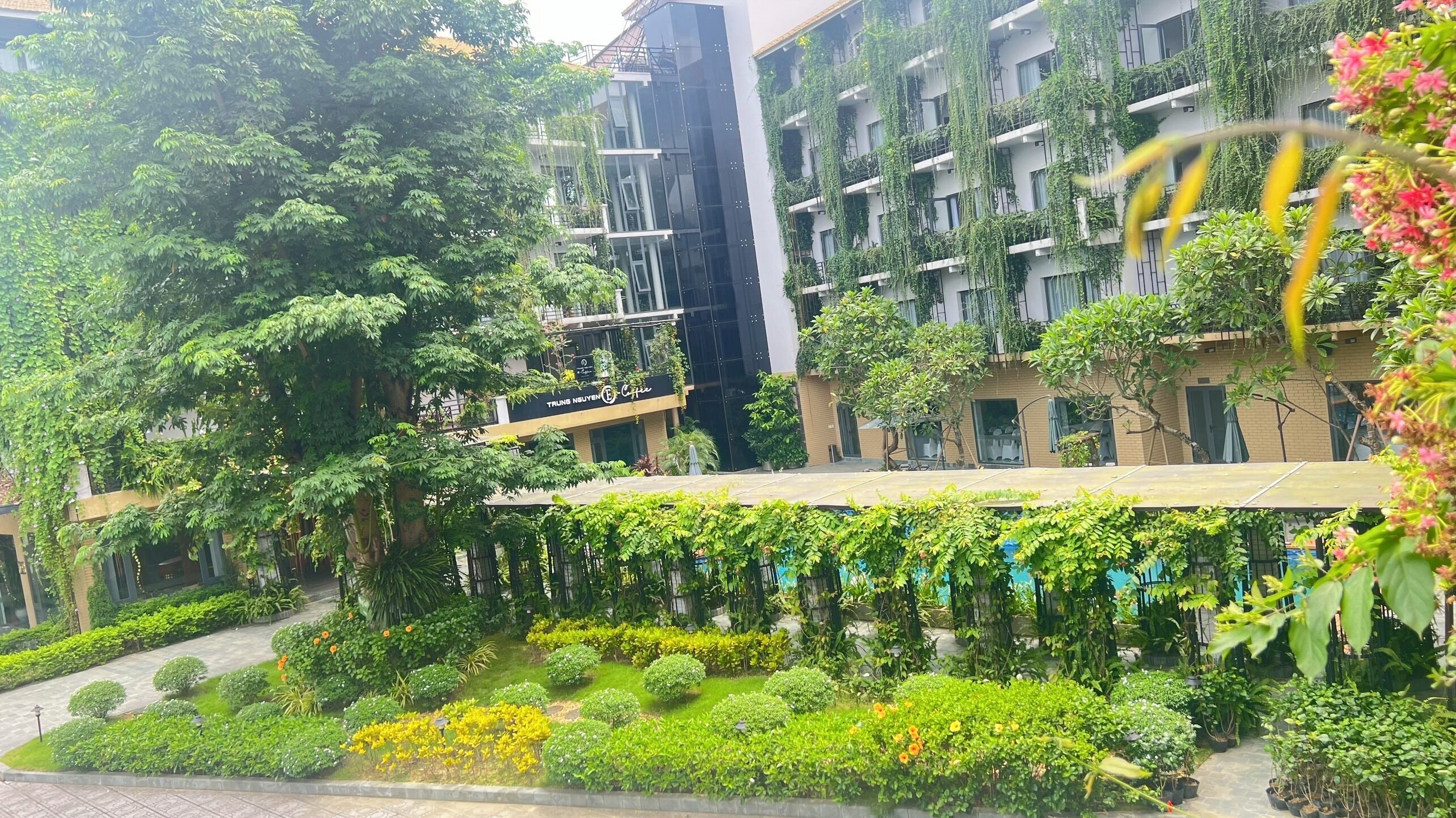 A multi-story building with balconies covered in greenery, a coffee shop on the ground floor, and a well-maintained garden with various plants and flowers.