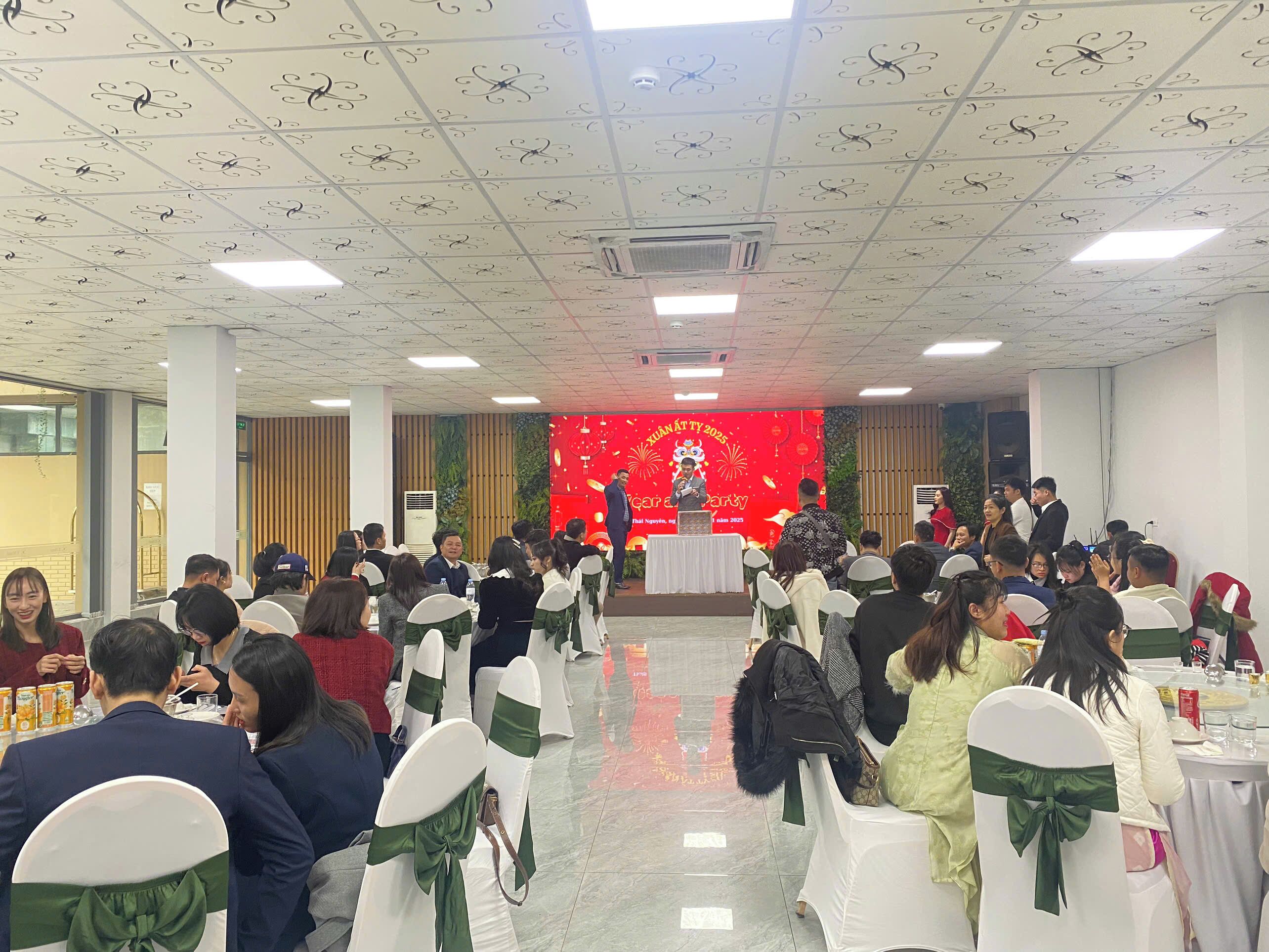 A conference room with white chairs, a stage with a red backdrop, and attendees seated facing the stage.