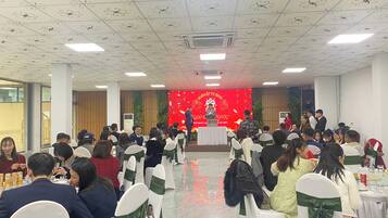A conference room with white chairs, a stage with a red backdrop, and attendees seated facing the stage.
