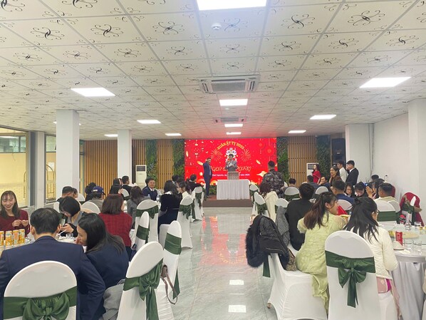 A conference room with white chairs, a stage with a red backdrop, and attendees seated facing the stage. - X Hotel Thai Nguyen (Phan Dinh Phung)