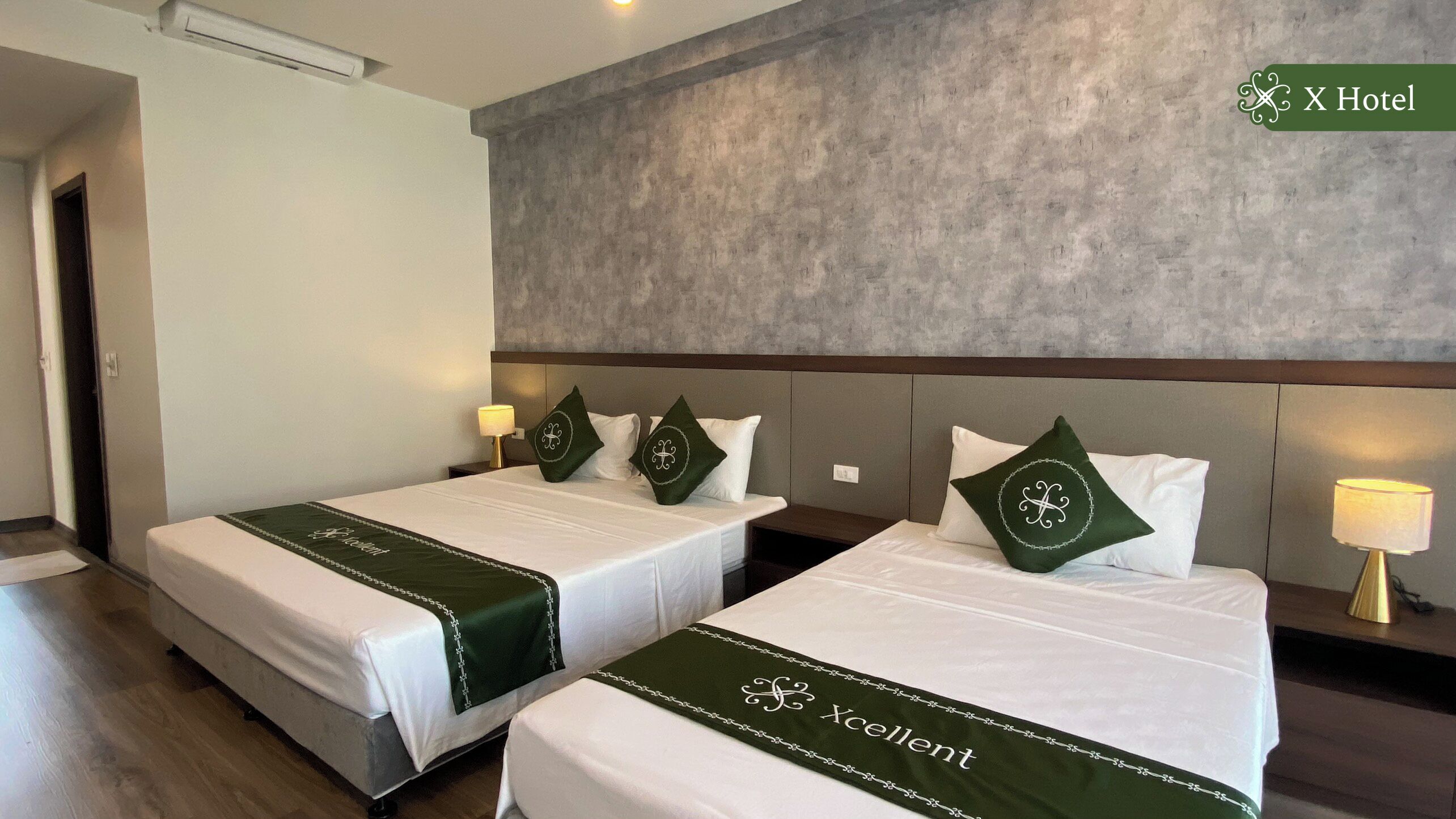 A hotel room with two beds, each with a green bedspread and matching pillows. The room has a concrete accent wall and a wooden headboard.