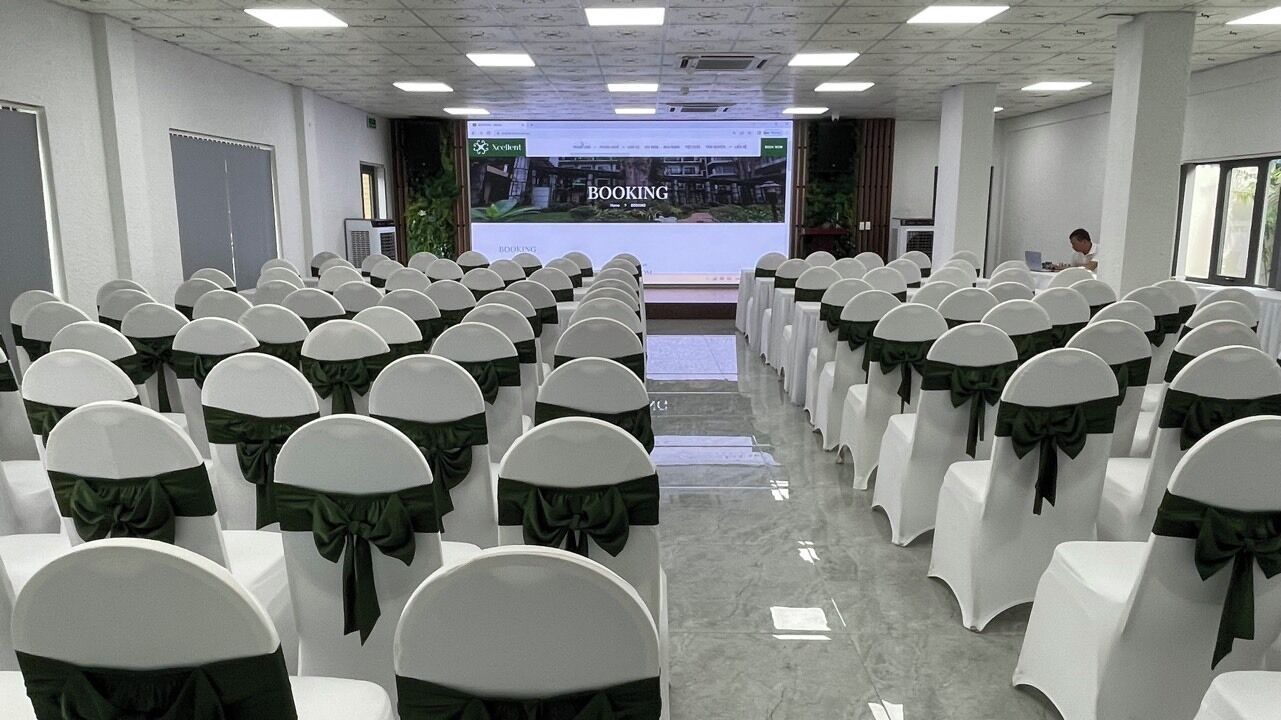 A conference room with rows of white chairs with green sashes, a podium at the front, and a large screen displaying a booking webpage.