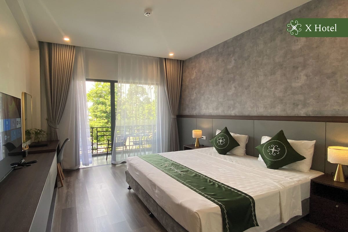 Ein Hotelzimmer mit einem großen Bett, einem Fernseher, einem Balkon mit Blick auf Bäume und einer grau strukturierten Wand.