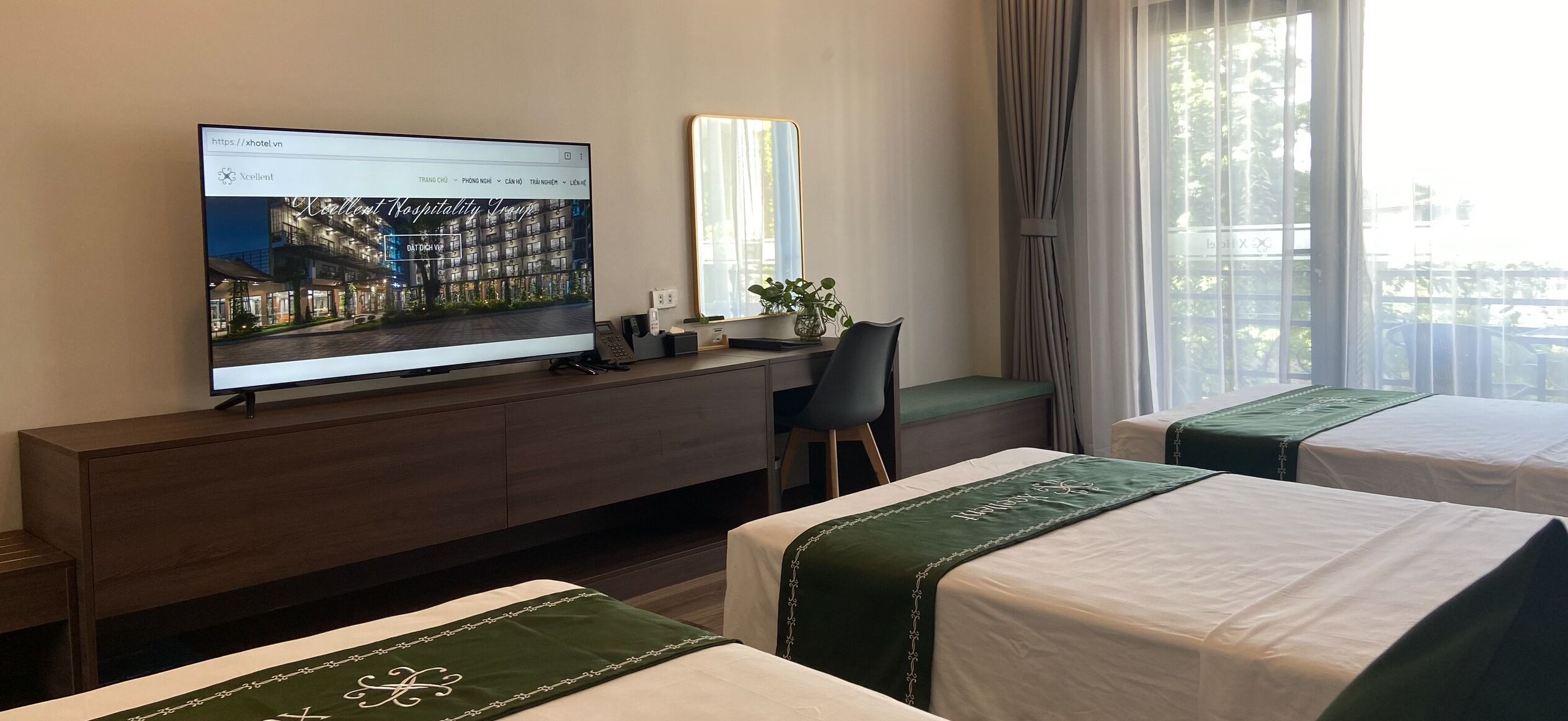 Ein Hotelzimmer mit Fernseher auf einer Holzkonsole, einem Schreibtisch mit Stuhl, zwei Betten mit grün-weißer Bettwäsche und einem großen Fenster mit Vorhängen.