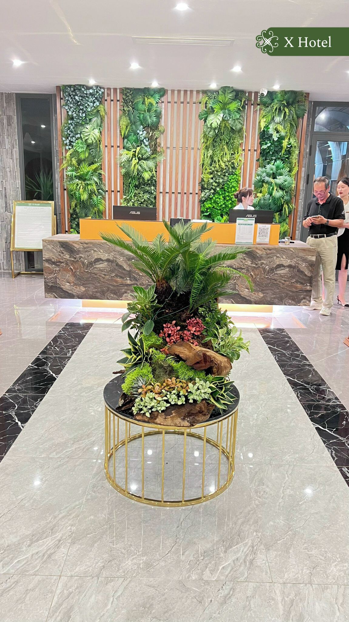A reception area with a marble floor, a central round table with a gold frame, and a backdrop of vertical greenery.