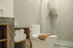 A modern bathroom with a toilet, a glass shower enclosure, and a shelf with neatly rolled towels. - MIMOYA STAY (Denpasar)