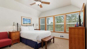 A bedroom with a bed, bedside tables, a red chair, a wooden bench, a ceiling fan, and large windows with blinds. - SeaEscape: Cute home, dog ok, near beach & pub! (Pacific City)
