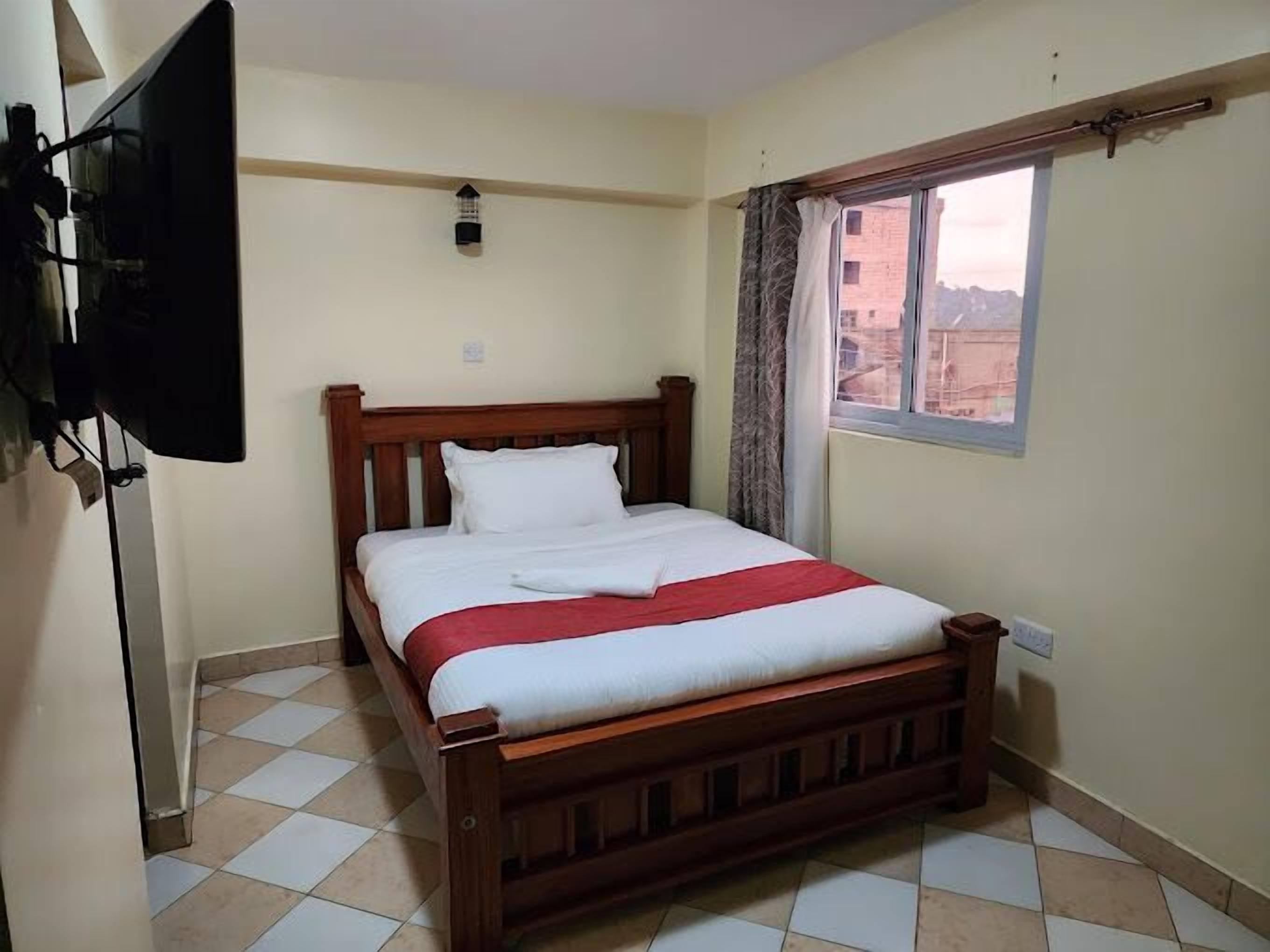 Quarto com cama de madeira, televisão de tela plana na parede, janela com cortinas e chão em azulejos.