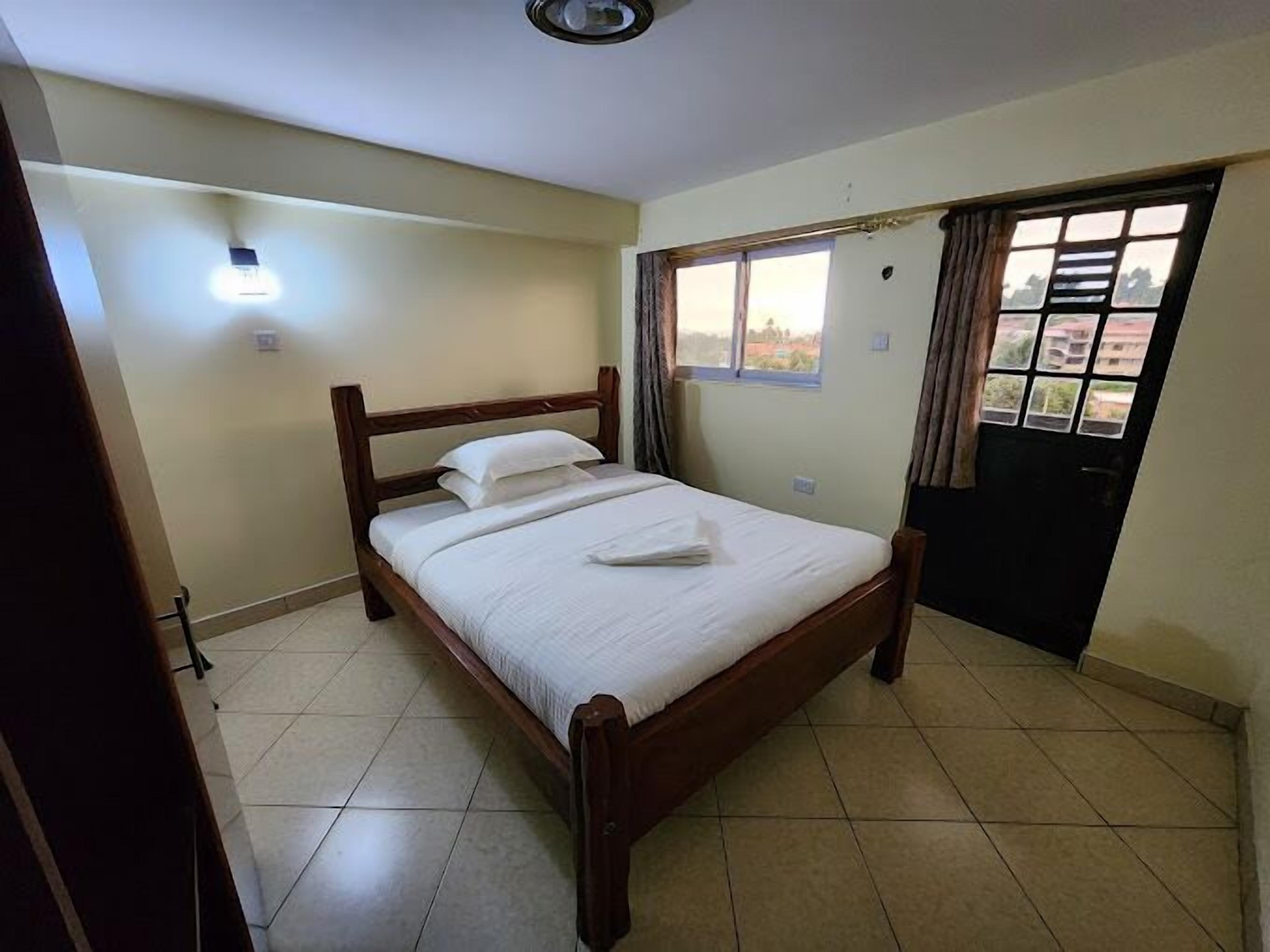 A bedroom with a wooden bed, a window with curtains, a door, and a wall-mounted light fixture.