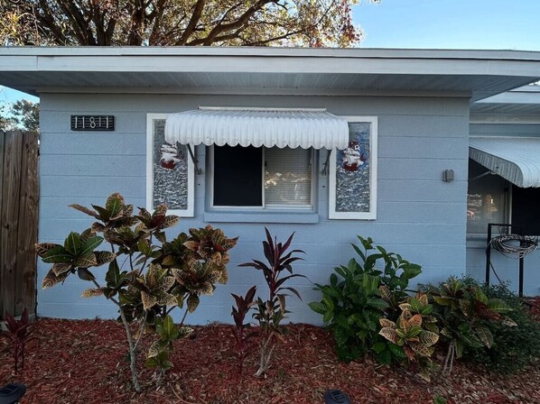 A single-story house with a light blue exterior, a white front door, and a small window with a white awning. There are various plants in the front yard, and a wooden fence is visible to the left.