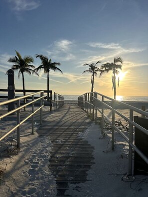 A wooden boardwalk leading to the beach with palm trees and a sunset in the background.