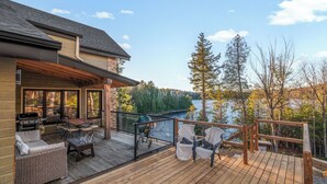 Una terraza de madera con mobiliario exterior, una parrilla y vistas a un lago rodeado de árboles.