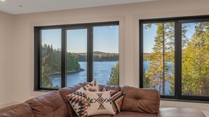 Habitación con sofá de cuero marrón y dos ventanas amplias que dan a vistas de un lago y un bosque.