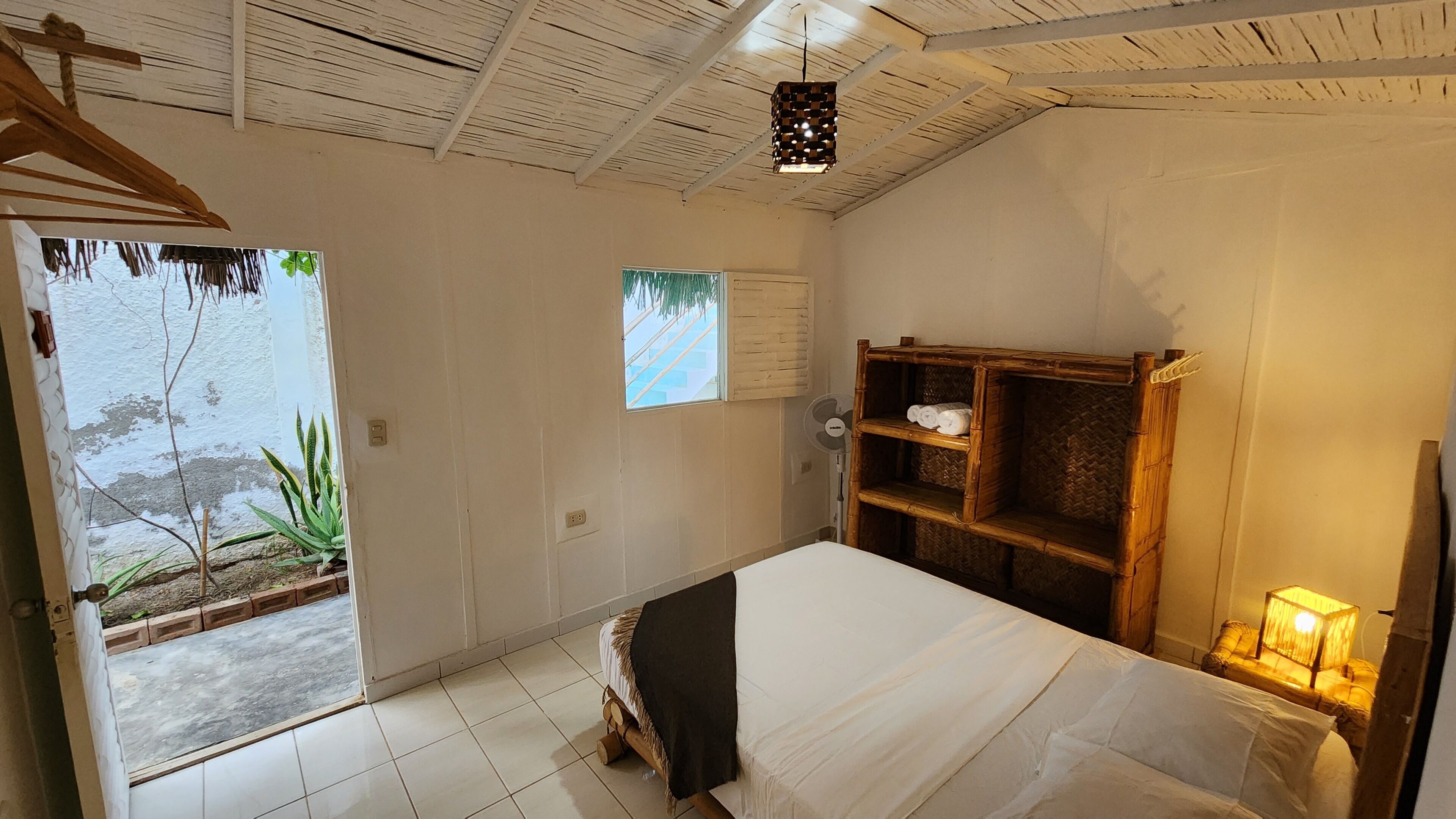 Una stanza con un letto, uno scaffale in legno, una ventola e una finestra.