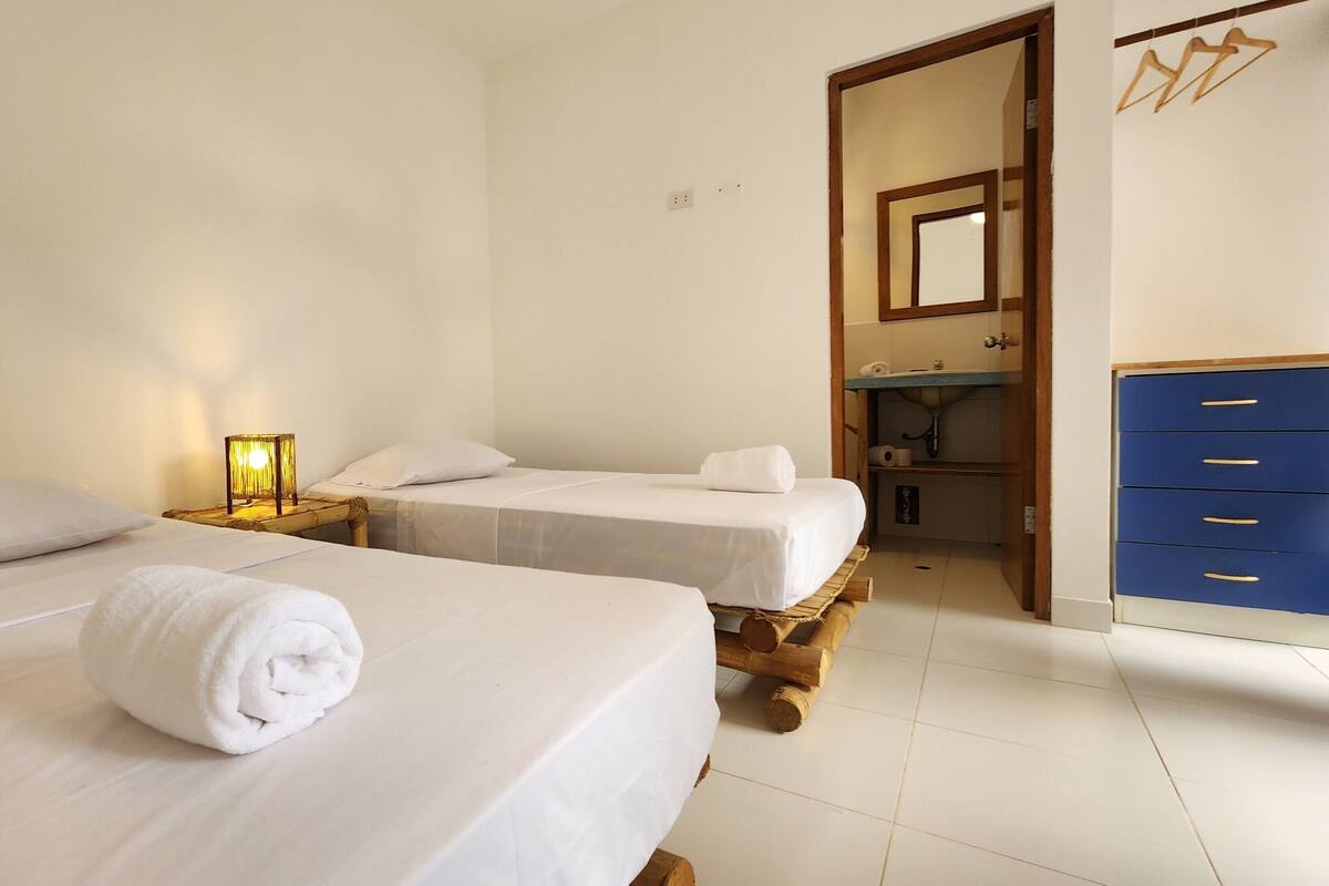 Une chambre d’hôtel avec deux lits, une table de chevet avec une lampe, et une salle de bain avec un miroir et des meubles bleus.