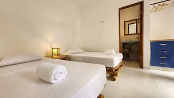 Una habitación de hotel con dos camas, una mesita de noche con lámpara, y un baño con espejo y armarios azules.