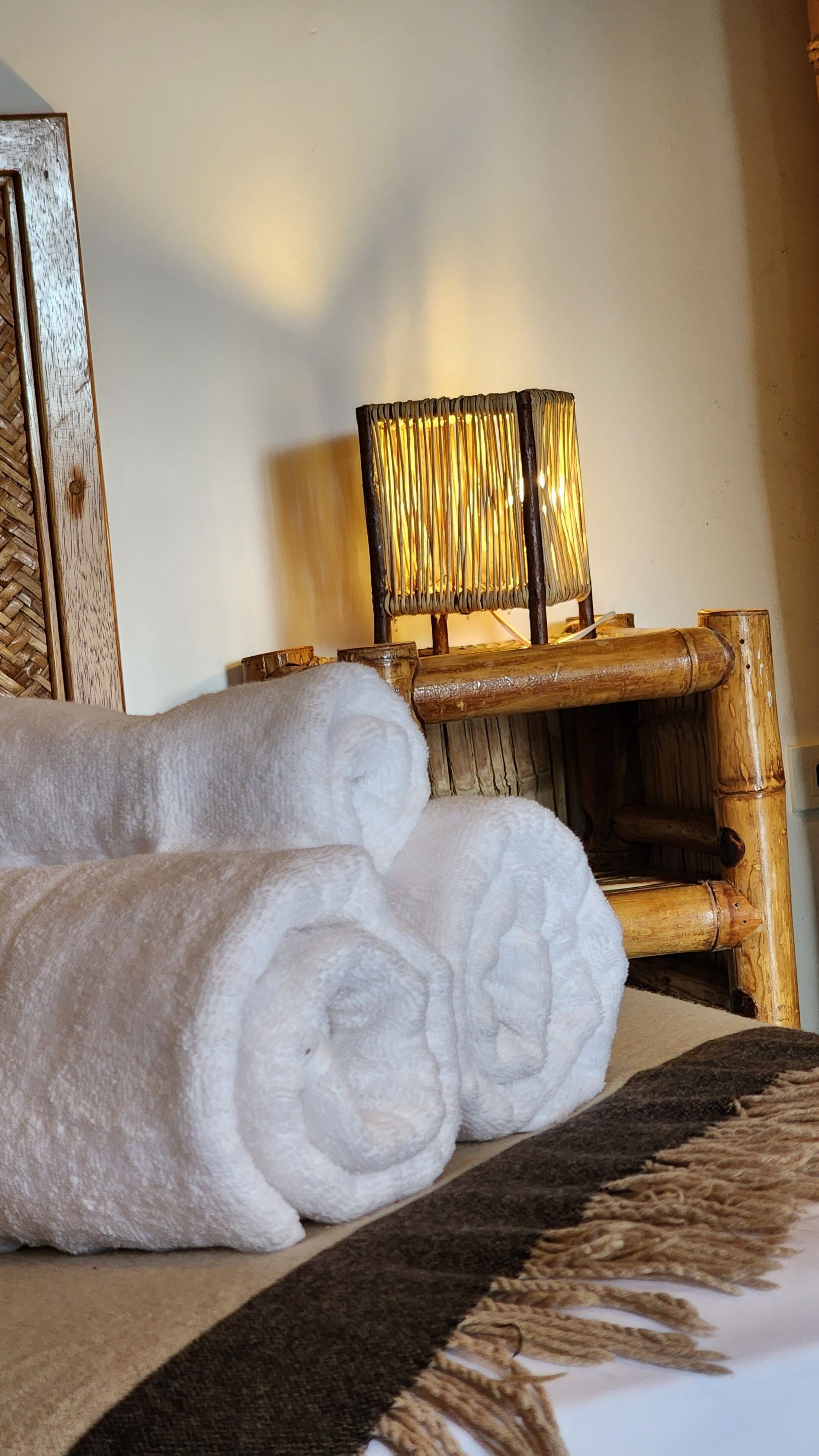 A bed with rolled white towels, a woven headboard, and a lamp with a golden shade.