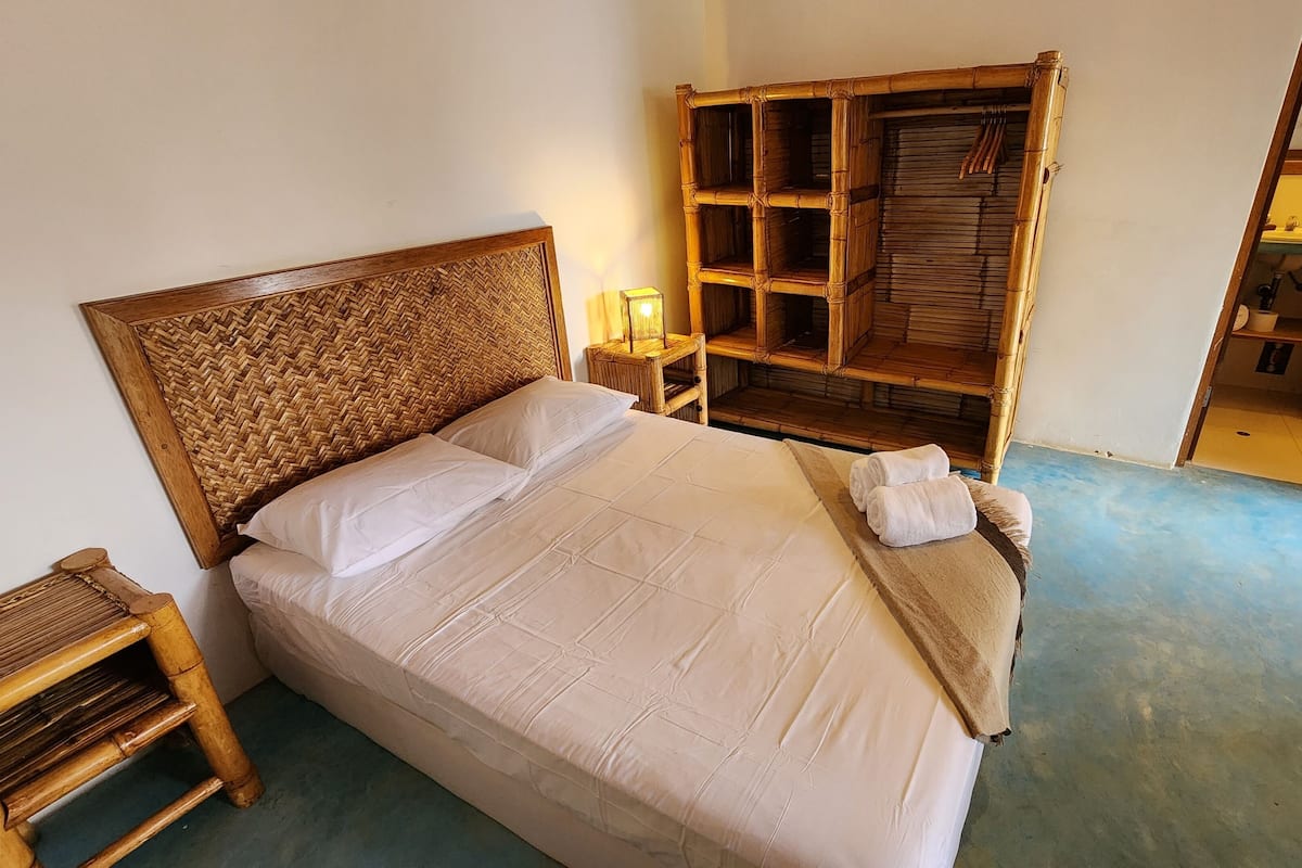 Un lit avec une tête de lit en osier, une table de chevet, une armoire en bois et une salle de bain visible à travers une porte ouverte.