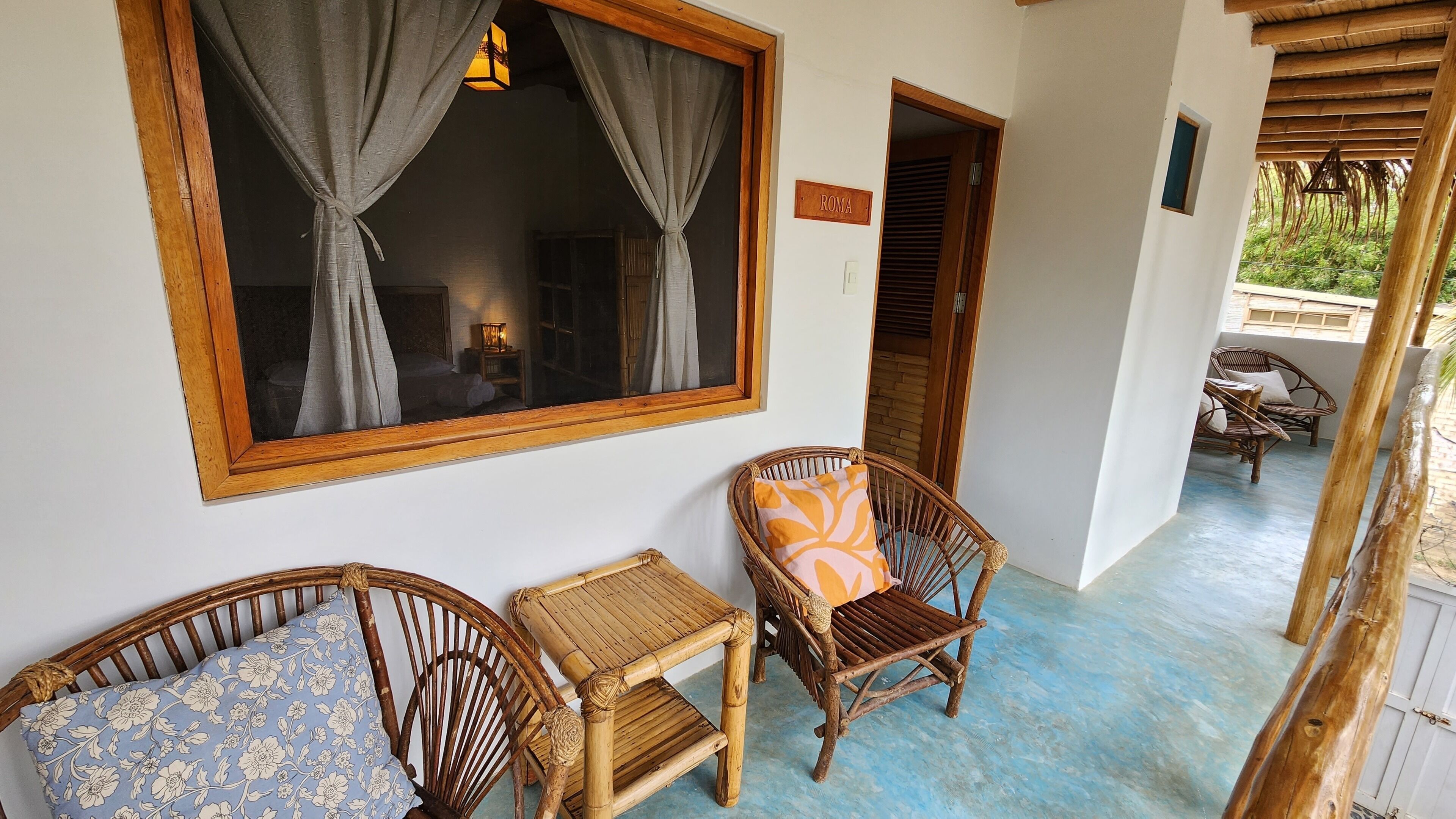 A room with a large window, a bed, a small table, and a wicker chair.
