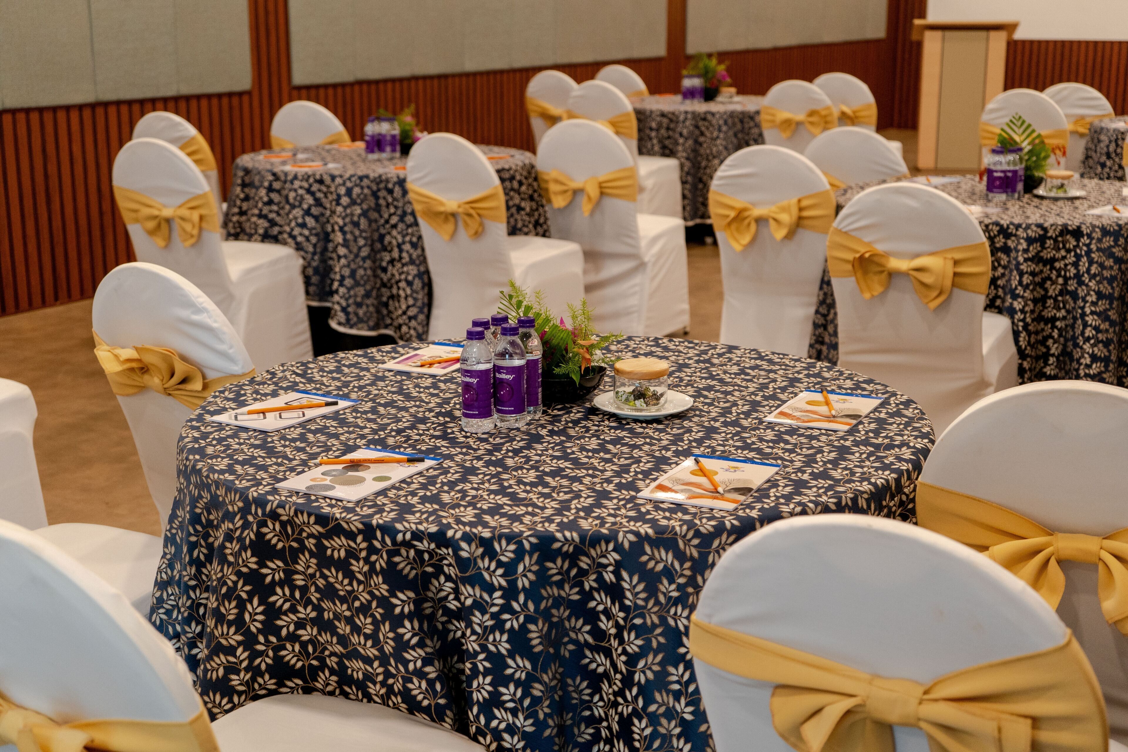 A conference room with round tables, white chairs with yellow sashes, floral tablecloths, and writing materials on the tables.