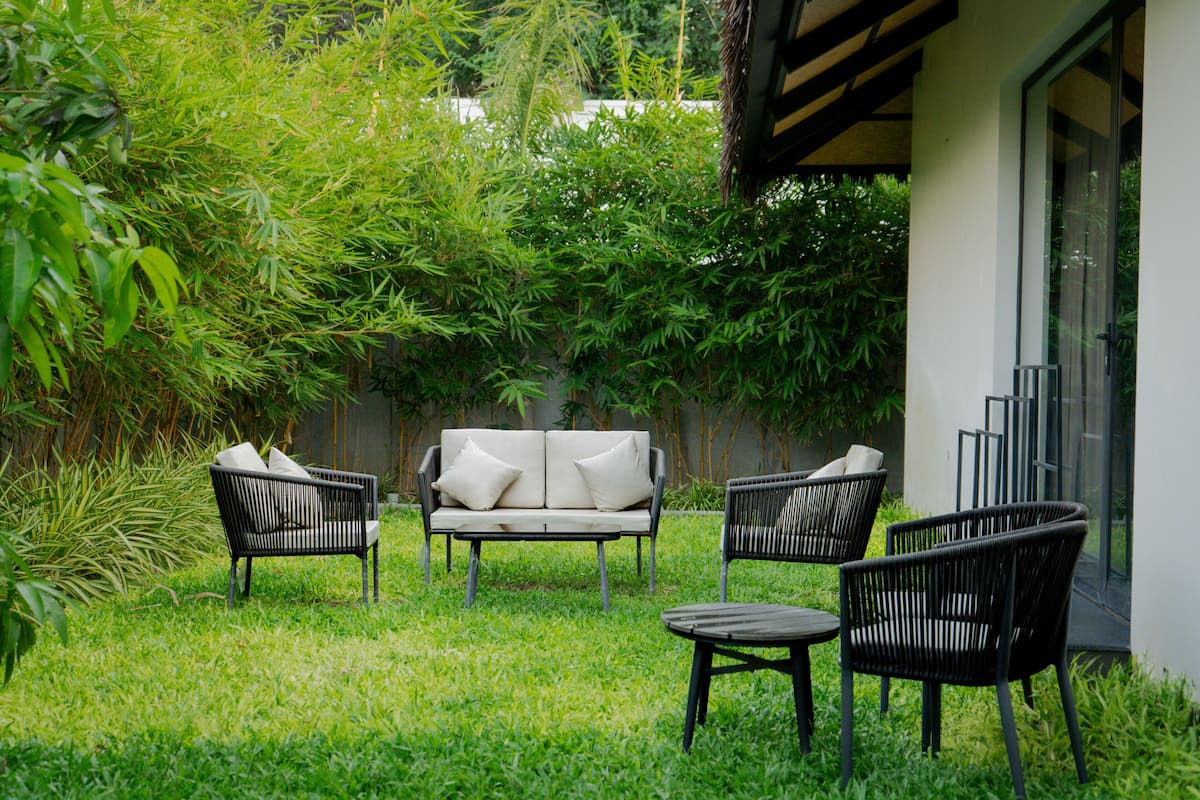 A patio area with a sofa, two chairs, and a table, surrounded by lush greenery and a building with a glass door.