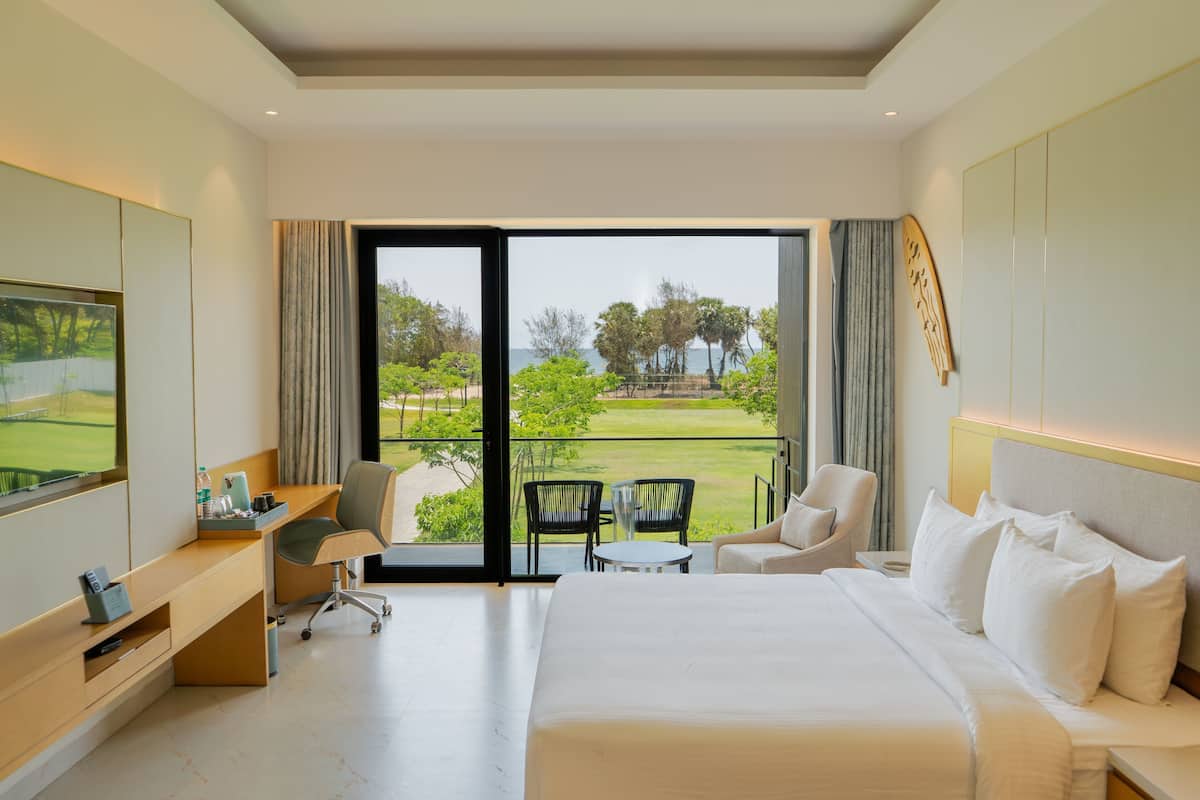 A hotel room with a large bed, a desk, a chair, and a balcony with a view of the greenery and water.
