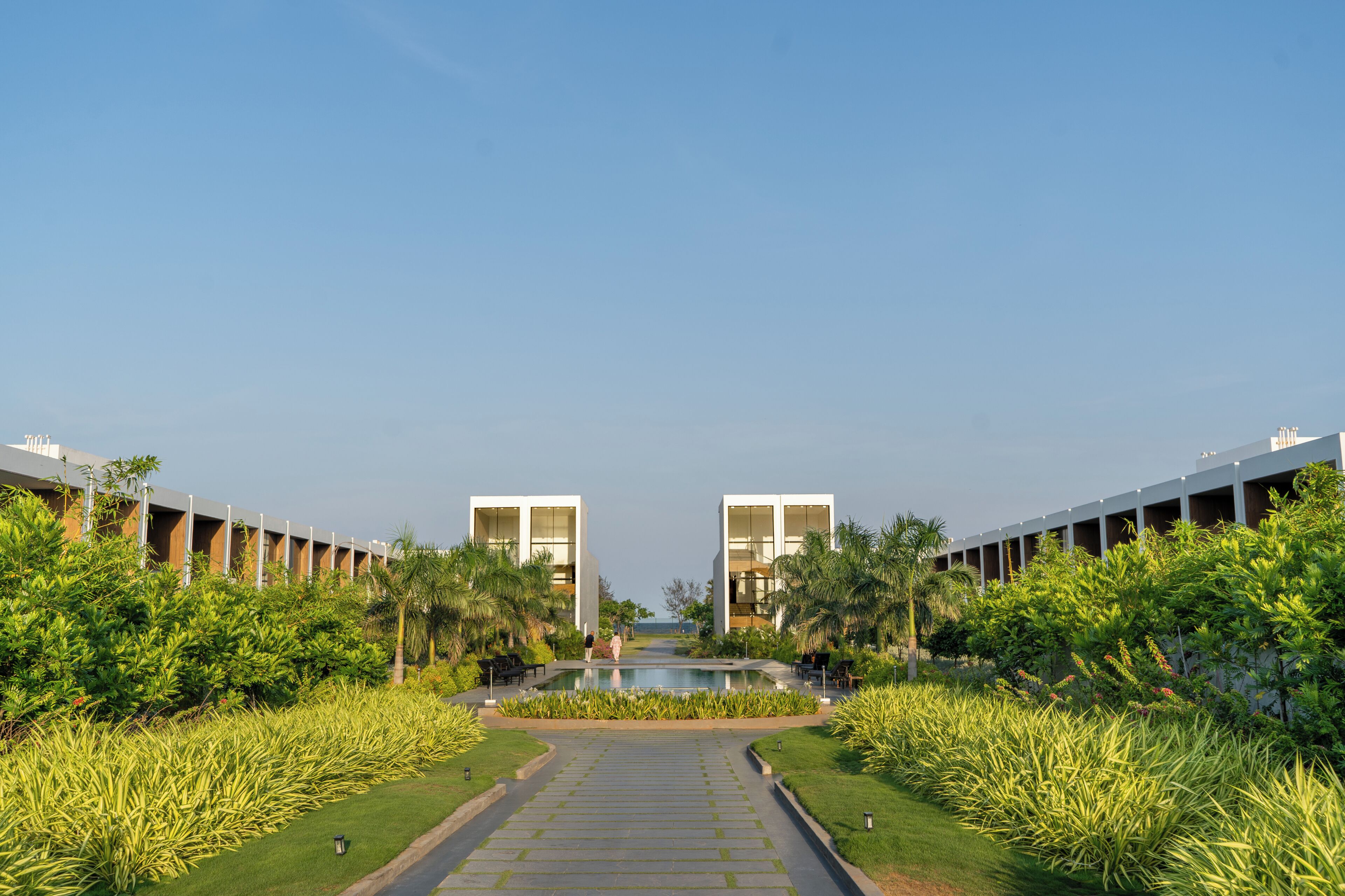 A pathway leading to a pool with modern buildings on either side, surrounded by greenery.