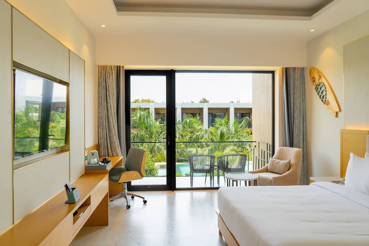 A hotel room with a bed, desk, chairs, a balcony with a pool view, and a surfboard decoration.