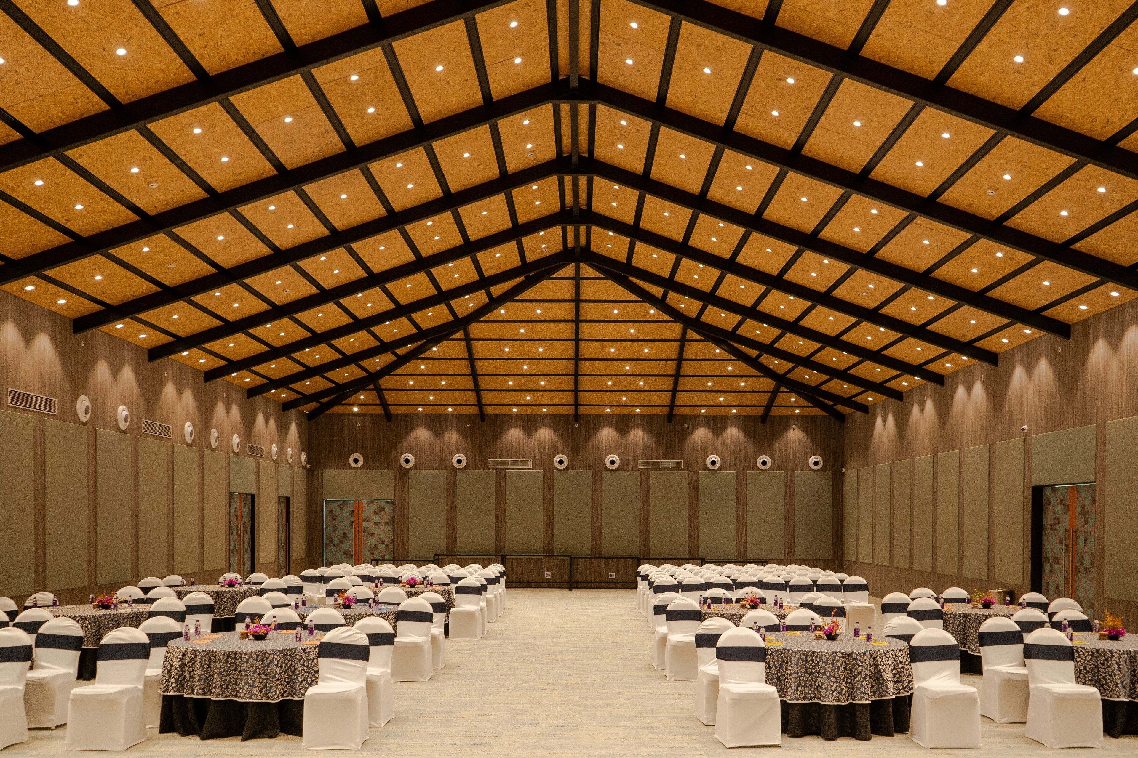 Un amplio salón de conferencias con filas de mesas y sillas, un techo alto con vigas de madera e iluminación empotrada.